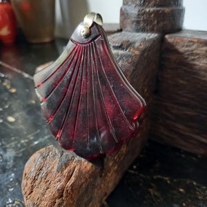 Pode incluir: Um pingente translúcido de cor vermelho-escuro em forma de leque ou concha, com sulcos verticais esculpidos. O pingente possui uma argola metálica prateada no topo para suspensão. O material apresenta uma aparência marmorizada escura.