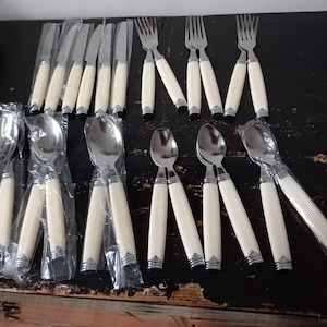Può includere: Una collezione di posate in acciaio inossidabile con manici a coste color avorio. Il set comprende coltelli, forchette e cucchiai, alcuni ancora nella loro confezione originale. Le posate sono disposte su una superficie di legno scuro.