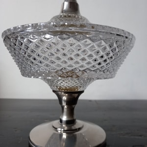 Puede incluir: Un platito de cristal transparente para dulces con una base y tapa de plata. El plato tiene un patrón de diamante y la base tiene un borde perlado.