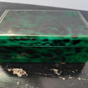 Könnte beinhalten: Eine rechteckige Box mit glänzendem, marmoriertem grün-schwarzem Finish. Die Box hat eine goldfarbene Umrandung um den Deckel. Die Box ist geschlossen und steht auf einer dunklen Oberfläche.