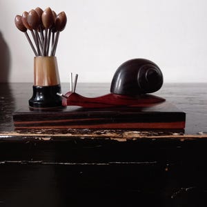 Könnte beinhalten: Eine Holzschneckenfigur mit dunkelbraunem Gehäuse und rotem Körper sitzt auf einem rechteckigen Sockel. Links befindet sich eine kleine Vase mit braunen tulpenförmigen Aufsätzen und Metallstielen. Die Objekte befinden sich auf einer dunklen Oberfläche.