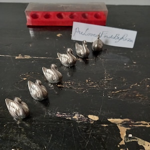May include: A collection of small, silver-colored swan figurines. The swans are arranged on a dark, distressed surface. A clear plastic container with a red base is visible in the background, along with a handwritten note that reads "PreLoved Finds By Roza."