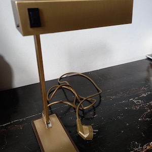 May include: A vintage brass desk lamp with a rectangular shade and a rectangular base. The lamp has a black on/off switch and a cord with a plug. The lamp is on a dark surface.