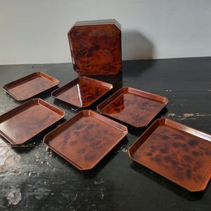 Puede incluir: Un juego de seis bandejas rectangulares con un patrón brillante, marrón y moteado similar al carey, dispuestas alrededor de una caja cuadrada a juego con tapa. Cada bandeja presenta bordes biselados y un acabado liso y reflectante, mostrando una paleta de colores marrón oscuro y ámbar consistente en toda la colección.
