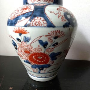 Può includere: Un vaso di ceramica bianca con corpo arrotondato e collo stretto. Il vaso è decorato con motivi floreali rossi e blu. La parte superiore del vaso ha una fascia blu con accenti floreali rossi. Il vaso poggia su una superficie scura.