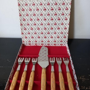Könnte beinhalten: Ein Vintage-Kuchenservierset in einer dekorativen Box. Das Set enthält einen Kuchenheber und sechs Gabeln. Die Griffe sind hellbeige mit einem bambusartigen Design. Die Box hat ein rotes Innenfutter und ein floral gemustertes Äußeres.