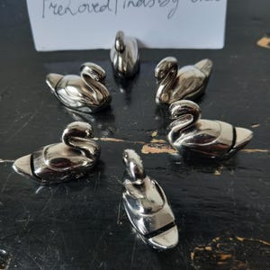 May include: A collection of small, silver-colored swan figurines arranged on a dark surface. The swans are detailed with curved necks and sleek bodies. A handwritten note above reads "PreLoved Finds By Roza".