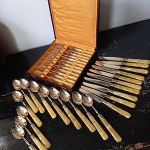 May include: A vintage silverware set with spoons, knives, and forks, arranged on a dark surface. The set includes multiple pieces with light-colored handles and silver-toned blades and bowls. The silverware is displayed both inside and outside of a box.