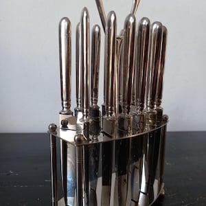 Op de afbeelding: Een set zilverkleurige metalen messen verticaal gerangschikt in een bijpassende ovale standaard. De standaard is voorzien van een centrale handgreep aan de bovenzijde en vier kleine bolvormige voetjes aan de basis. Elk mes heeft een glad handvat.