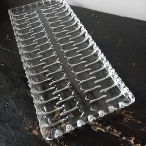 May include: A clear, rectangular glass serving tray with a textured, ridged design. The tray's edges have a scalloped pattern, and the overall aesthetic is vintage or retro. The glass reflects light, creating highlights and shadows.