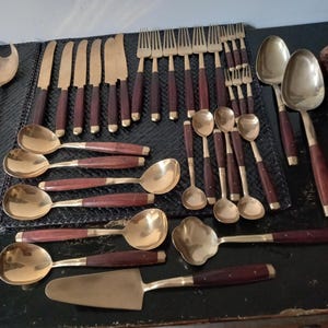 Può includere: Un set di posate vintage con teste color ottone e manici in legno scuro. Il set include coltelli, forchette, cucchiai e utensili da portata. La disposizione è su una superficie scura, mostrando il set da pranzo completo.