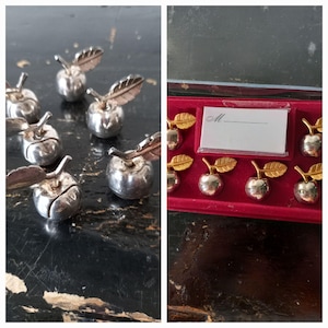 May include: A set of small silver-toned apple-shaped place card holders with stems and leaves. Some are displayed loose, while others are arranged in a red velvet-lined box alongside a white rectangular card featuring the letter M and a line.