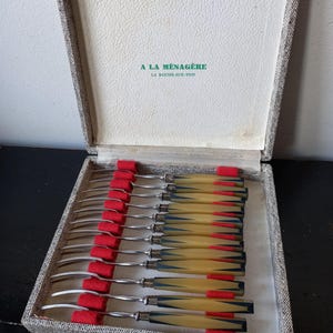 Puede incluir: Un juego de 12 tenedores de plata con mangos amarillos y rojos en una caja de tela gris. La caja tiene una etiqueta que dice "A La Ménagère La Roche-sur-Yon".