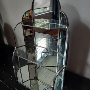 Op de afbeelding: Een displayplank met meerdere niveaus, opgebouwd uit spiegelglaspanelen die bijeengehouden worden door een donker metalen frame. De unit heeft een boogvormige bovenkant, twee horizontale planken en een semi-hexagonale basis.