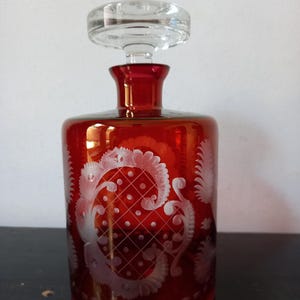 May include: A cylindrical red glass decanter featuring a clear glass stopper and intricate etched white floral and lattice patterns around the body. The vessel has a short neck and a flat base, showcasing a vibrant crimson hue throughout the glass.