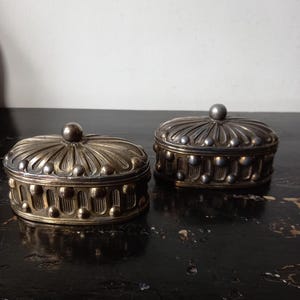 May include: Two antique oval-shaped metal boxes with lids. The boxes have a detailed design with a mix of gold and silver tones. Each box features a small, round knob on top of the lid. The boxes are placed on a dark, textured surface.