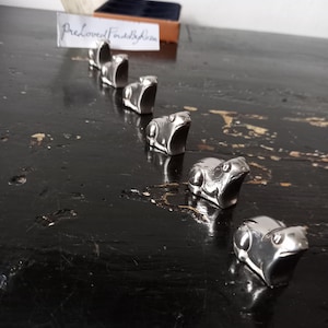 May include: Six silver frog ornaments arranged in a diagonal line on a black, distressed surface. The frog ornaments are small and detailed. A sign with handwritten text is in the background.