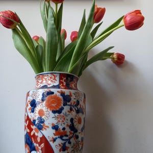 Puede incluir: Un jarrón de cerámica adornado lleno de tulipanes rojos. El jarrón presenta un detallado patrón floral en tonos azules, naranjas y blancos. Los tulipanes tienen tallos y hojas verdes, creando un contraste con el diseño del jarrón.