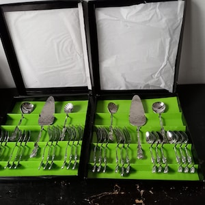 Peut inclure: Deux ensembles de couverts argentés dans des boîtes noires avec des inserts verts. Chaque ensemble comprend des fourchettes, des cuillères, un serveur à gâteau et d'autres ustensiles. Les boîtes sont ouvertes, révélant l'argenterie bien rangée.