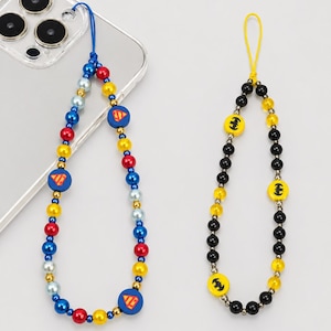 Op de afbeelding: Twee telefoonbedels met lussen. De ene heeft blauwe, rode, gele en lichtblauwe kralen met ronde Superman-logo bedels aan een blauw koord. De andere heeft zwarte en gele kralen met ronde Batman-logo bedels aan een geel koord.