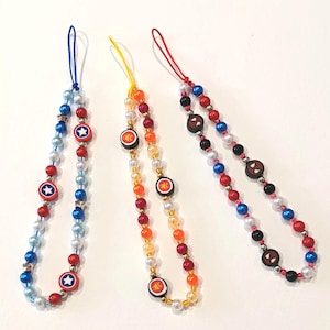 May include: Three beaded phone charms with superhero designs. One features a Captain America shield, another has a circular design, and the third showcases a Spider-Man face. The beads are in various colours, including red, blue, white, and black.