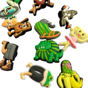 May include: A selection of vibrant shoe charms in diverse designs. The charms feature a green owl, a brown character, a green and orange shoe, a green alligator, a ballerina, a monkey in a banana, and others. These charms are for shoe decoration.