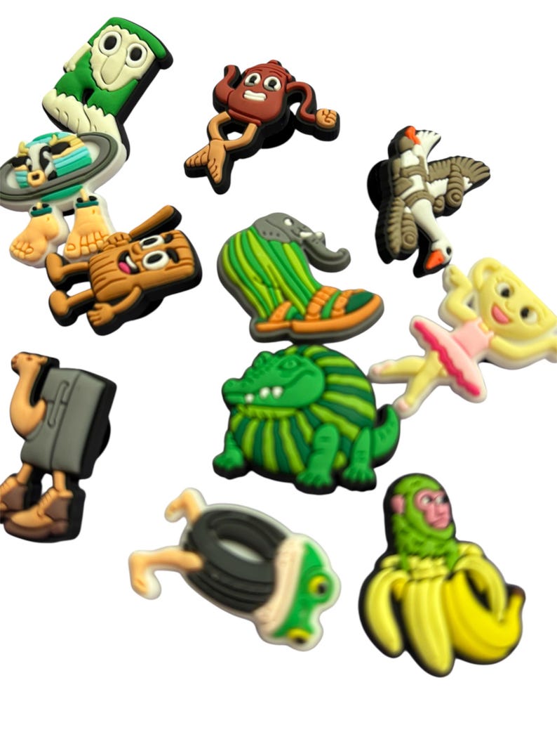May include: A selection of vibrant shoe charms in diverse designs. The charms feature a green owl, a brown character, a green and orange shoe, a green alligator, a ballerina, a monkey in a banana, and others. These charms are for shoe decoration.
