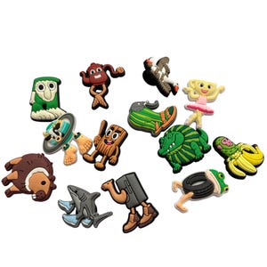 May include: A selection of vibrant shoe charms featuring cartoon characters. The charms depict a green monster, a shark, a ballerina, a bear, and other playful designs. These charms are made to be attached to footwear.