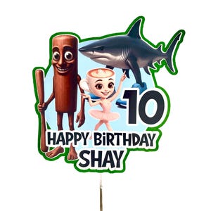 May include: A vibrant cake topper featuring a cartoon shark, a ballerina in a coffee cup, and a wooden figure with a bat. The text "HAPPY BIRTHDAY SHAY" and the number "10" are displayed on the topper.