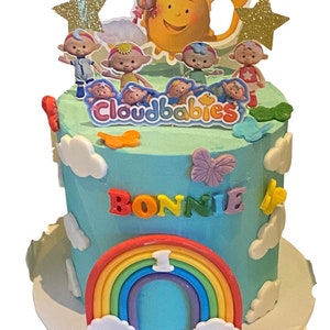 May include: A blue and white birthday cake decorated with a rainbow, clouds, and the characters from the show "Cloudbabies". The cake is topped with a sun and stars. The cake says "Bonnie" and "1" on it.