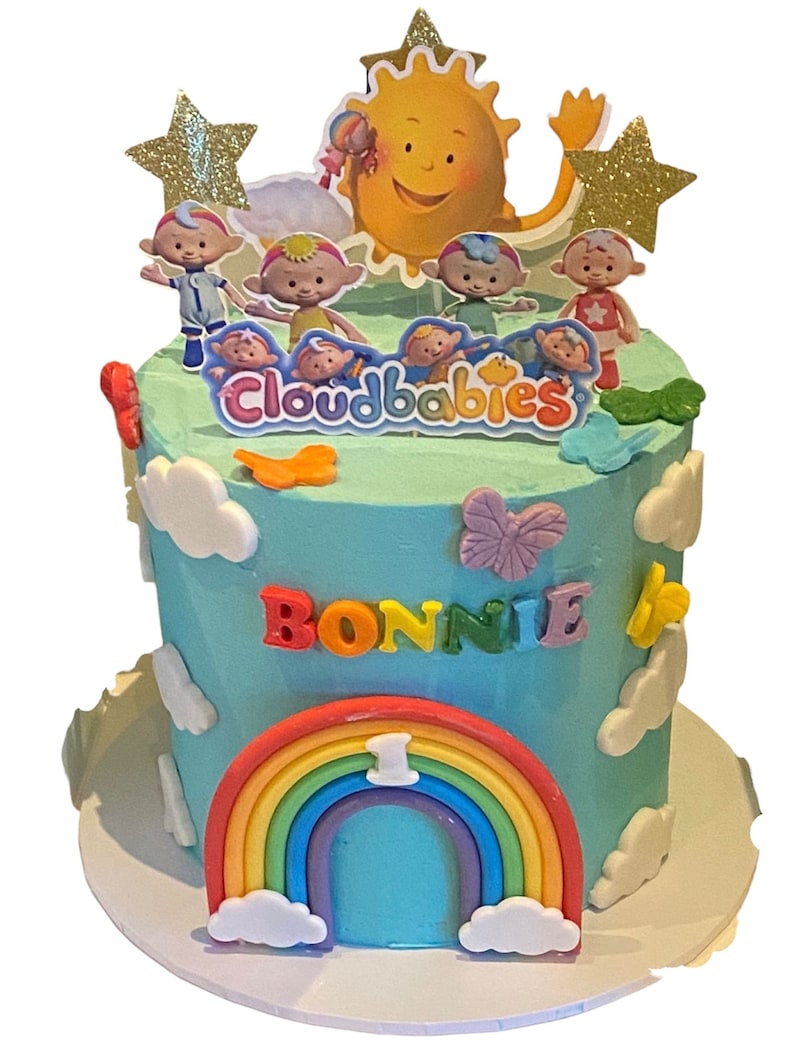 May include: A blue and white birthday cake decorated with a rainbow, clouds, and the characters from the show "Cloudbabies". The cake is topped with a sun and stars. The cake says "Bonnie" and "1" on it.