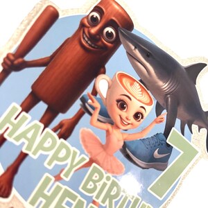May include: A vibrant birthday cake topper with cartoon characters. Includes a shark, a coffee cup ballerina, and a cinnamon stick with a baseball bat. The text reads "HAPPY BIRTHDAY HENDRI" and the number "7".