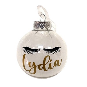 May include: Clear glass Christmas bauble featuring black eyelashes and the name "Lydia" in gold script. It has a silver cap and a white ribbon. A decorative festive ornament for the holiday season.