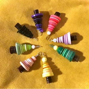 May include: A set of eight colourful button Christmas tree decorations. Each decoration is made of a stack of buttons in various colours and patterns, including purple, pink, green, and yellow. The decorations are strung on silver chains and have a brown button at the bottom.
