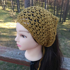May include: Mustard yellow crochet head covering with a net-like design. The head covering is tied at the back and is displayed on a mannequin head with dark hair. The background is a blurred outdoor scene with trees.