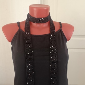 May include: A black crocheted scarf featuring a choker-style neck wrap and two long, dangling ends. The fabric has a textured, open-weave pattern and is embellished with small, reflective silver sequins scattered throughout the entire length.