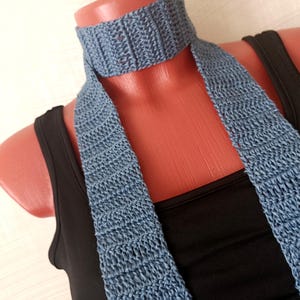 Könnte beinhalten: Ein staubblauer Häkel-Choker-Schal mit einem strukturierten Rippenmuster. Das Accessoire liegt eng am Hals an und hat zwei lange, flache, vertikale Bahnen, die über einem schlichten schwarzen ärmellosen Oberteil nach unten verlaufen.