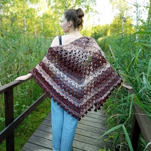 May include: A triangular crochet shawl featuring an intricate openwork pattern in variegated shades of deep burgundy, dark brown, and cream. The garment has a scalloped edge with small decorative tassels along the bottom border and drapes over the shoulders.