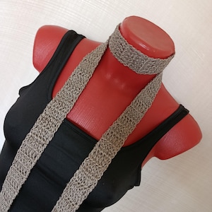 May include: A beige crocheted choker necklace with two long, vertical crocheted strips extending downwards, worn over a plain black sleeveless top. The crochet work features a consistent, textured stitch pattern throughout the entire accessory.