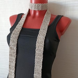 May include: A beige knitted scarf and choker set displayed on a mannequin. The scarf is long and rectangular, with a textured, open weave. The choker is a matching band. The mannequin is wearing a black tank top.