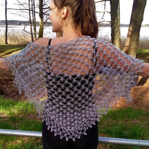 Puede incluir: Un chal de ganchillo en hilo metálico plateado con un patrón de celosía calado y pequeñas lentejuelas brillantes integradas. La prenda triangular se coloca sobre los hombros, mostrando una construcción ligera y aireada con un diseño delicado y repetitivo en forma de nudo en toda la pieza.