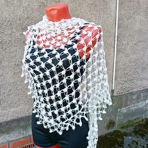 May include: A cream-colored, open-weave shawl with a delicate, knotted pattern. The shawl is draped over a black top on a mannequin. The design features small, decorative bobbles along the edges, adding texture and visual interest to the garment.