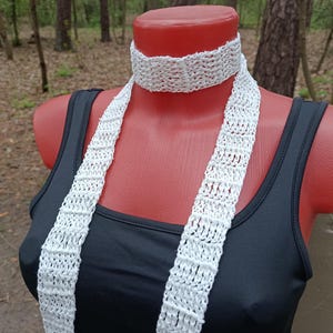 Peut inclure: Une écharpe blanche à tissage ouvert est drapée autour du cou d'un mannequin. L'écharpe est faite d'une matière texturée, peut-être crochetée ou tricotée. Le mannequin porte un débardeur noir. L'arrière-plan est une scène extérieure floue avec des arbres.
