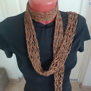 May include: A long, brown macrame scarf featuring an open, knotted net pattern. The scarf is draped around the neck of a mannequin wearing a plain black short-sleeved shirt. The intricate, hand-tied cord design creates a textured, airy appearance.