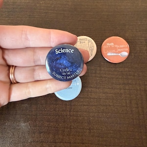 May include: A collection of round buttons with educational themes. One button features the word "Science" with "Cycle 2" and "Subject Master" text. Other buttons include "History 800" and "Math" with additional text. The buttons are in various colors.