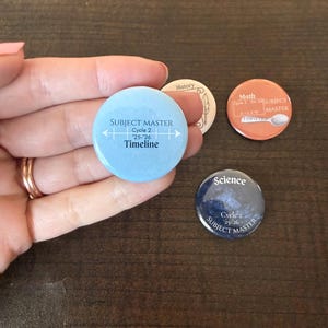 Può includere: Una collezione di bottoni rotondi a tema educativo. Un bottone è azzurro con "SUBJECT MASTER Cycle 2 '25-'26 Timeline". Altri bottoni includono "History", "Math" e "Science" con testo correlato.