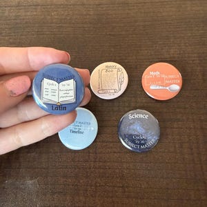 May include: A collection of five round buttons with educational themes. The buttons feature the words "Latin", "History", "Math", and "Science", along with the text "Subject Master" and "Cycle 2". The buttons are in various colors, including blue, orange, and white.