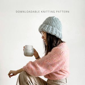 May include: A person wearing a light blue knitted beanie, a pink fuzzy sweater, and beige pants sits on a wooden stool holding a ceramic mug.