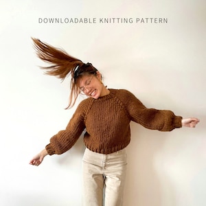 May include: A woman wearing a brown knitted sweater with a high neckline and long sleeves. The sweater is made with chunky yarn and has a relaxed fit. The text "Start Here Top Down" and "By Brenda Lam" is visible at the bottom of the image.