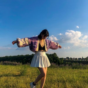 Peut inclure: Une femme portant une jupe pliss&eacute;e blanche, un soutien-gorge noir et un cardigan en tricot violet et blanc &agrave; motif floral. Elle se tient dans un champ d'herbe verte avec un ciel bleu en arri&egrave;re-plan.